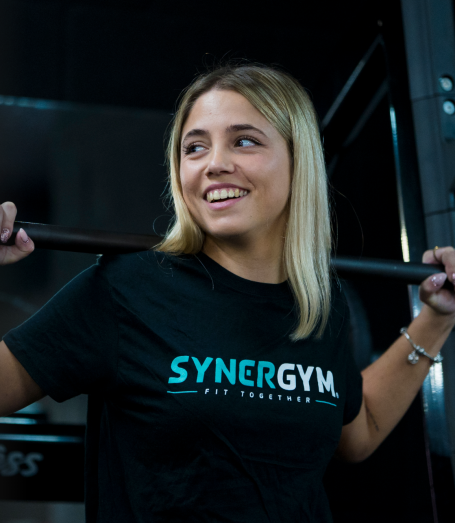 Entrenamiento en SYNERGYM Joven sonriendo mientras sostiene una barra en un gimnasio. Lleva una camiseta negra.