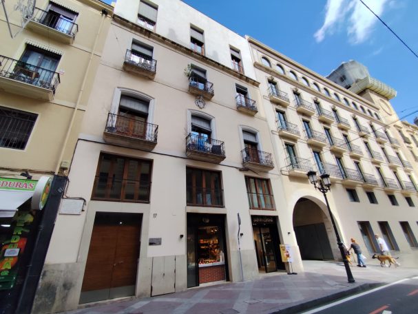Intermediación en la compraventa de un edificio en el centro de Alicante, destinado a apartamentos turísticos Edificio de varios pisos con balcones y comercios en la planta baja, cielo azul.