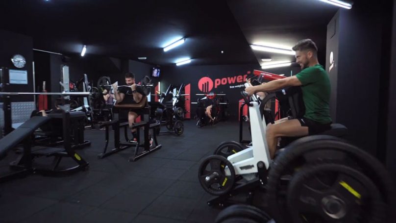 Instalaciones SYNERGYM Gimnasio moderno con personas entrenando en diferentes máquinas y áreas de ejercicio.
